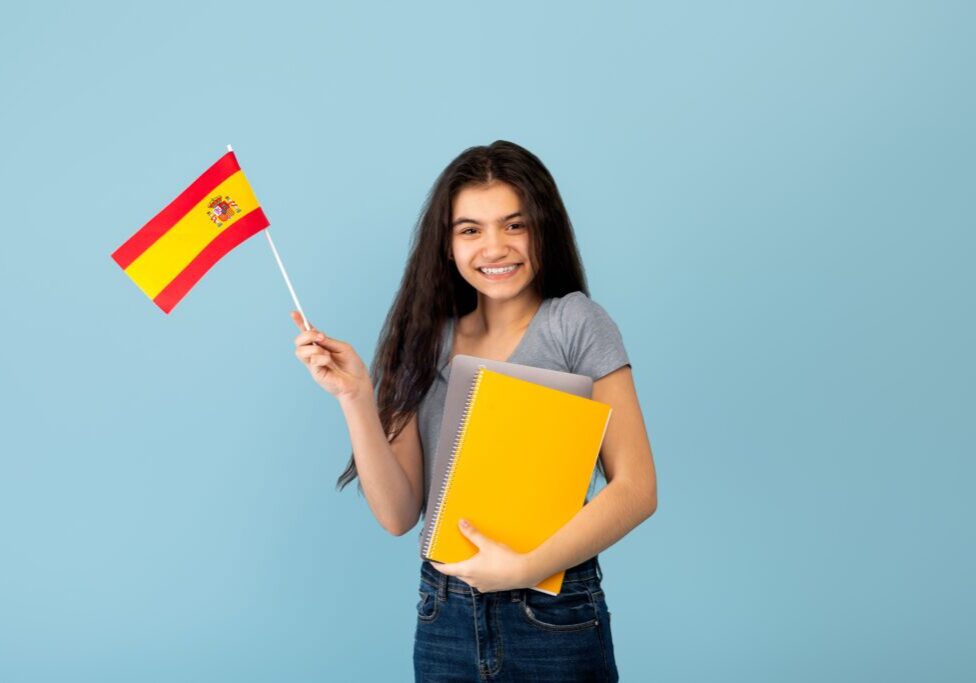 Cheerful Indian teen exchange student with notebook and laptop pc holding flag of Spain on blue studio background. Happy adolescent girl studying Spanish abroad online