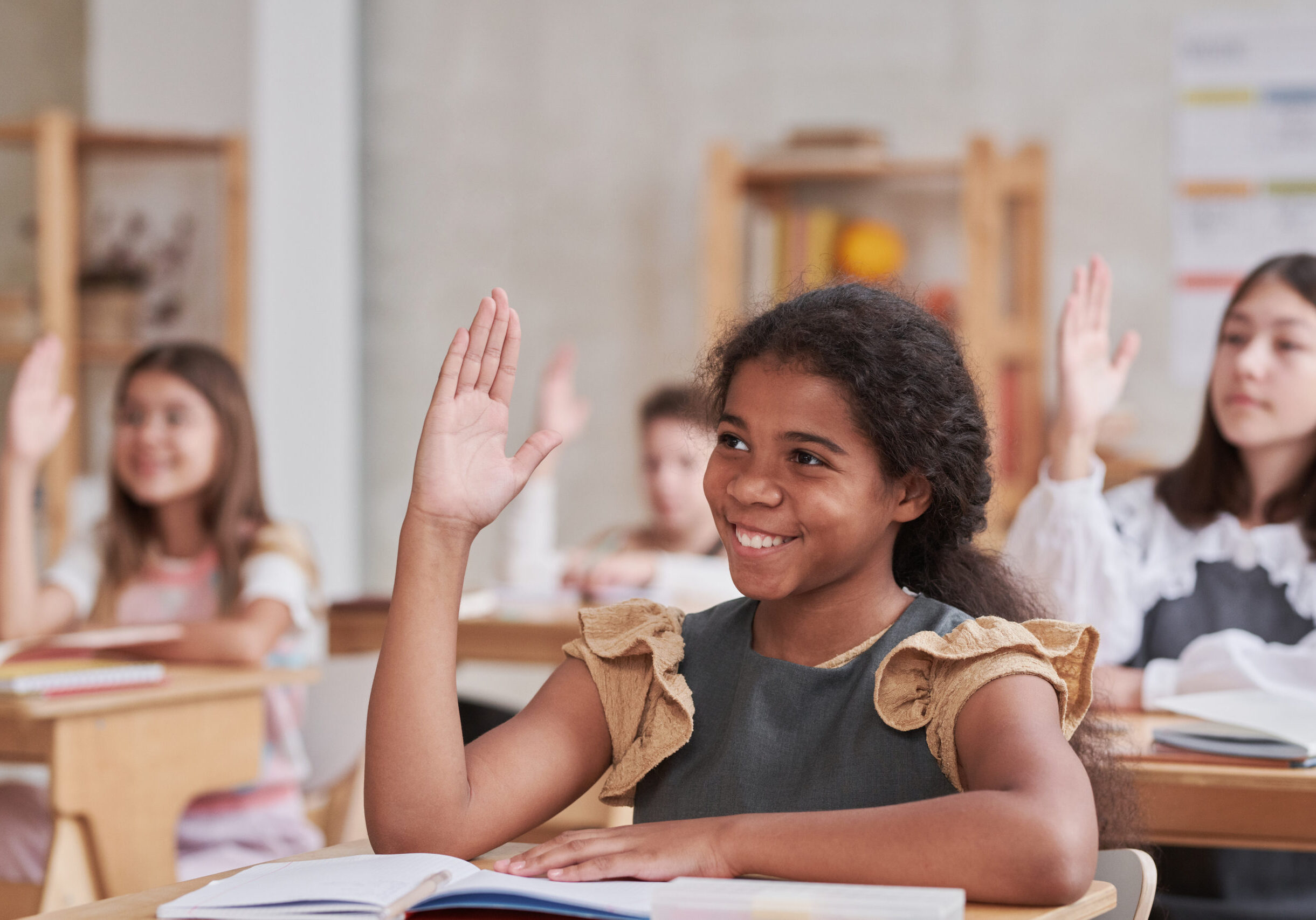 Portrait of smiling African-American schoolgirl raising hand while sitting at desk in classroom, copy space